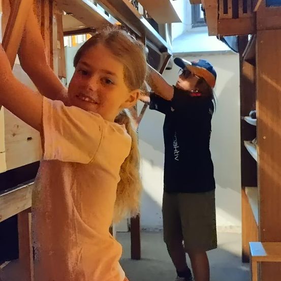 Ein junges Mädchen mit blonden Haaren klettert auf eine Holzkonstruktion, während ein Erwachsener mit Hut und Brille daran arbeitet.
