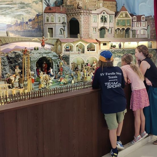 Drei Kinder und ein Erwachsener betrachten eine Miniatur-Krippenszene mit einem detaillierten Stadthintergrund. Der Junge trägt eine blaue Kappe und ein blaues Shirt mit weißem Text.