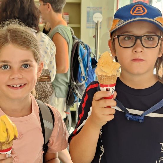 Zwei Kinder, ein Mädchen und ein Junge, halten Eisbecher in der Hand. Das Mädchen lächelt und der Junge schaut in die Kamera. Sie stehen an einem öffentlichen Ort.