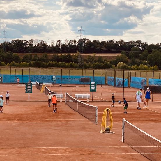 Eine Gruppe von Menschen spielt Tennis auf einem Sandplatz. Einige stehen in der Nähe der Netze, während andere herumlaufen. Der Platz ist von einem Zaun umgeben, und im Hintergrund befinden sich Bäume und Stromleitungen.