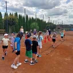 Eine Gruppe von Kindern nimmt auf einem Tennisplatz an einer Sportaktivität teil, wobei einige laufen und andere herumstehen. Sie tragen Mützen und Turnschuhe. Im Hintergrund befindet sich ein Zaun und Bäume.