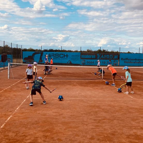 Eine Gruppe von Kindern spielt Tennis auf einem Sandplatz, mit einem Netz, das den Platz teilt. Einige halten Schläger, während andere Bälle aufheben. Es gibt Werbebanner am Zaun.
