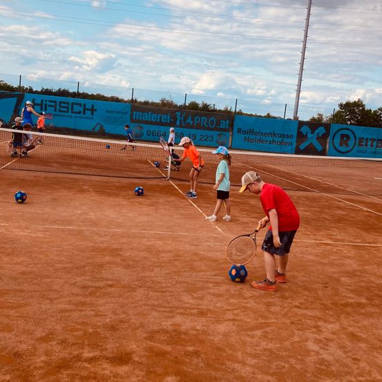 Eine Gruppe von Kindern spielt Tennis auf einem Sandplatz mit Netz unter einem teilweise bewölkten Himmel. Einige halten Schläger, andere sitzen an den Seitenlinien.