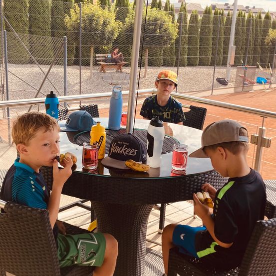 Drei Jungen sitzen an einem Tisch draußen, essen Snacks und trinken aus Bechern. Ein Junge trägt eine Yankees-Kappe und ein anderer eine Mütze. Ein Tennisplatz ist im Hintergrund.