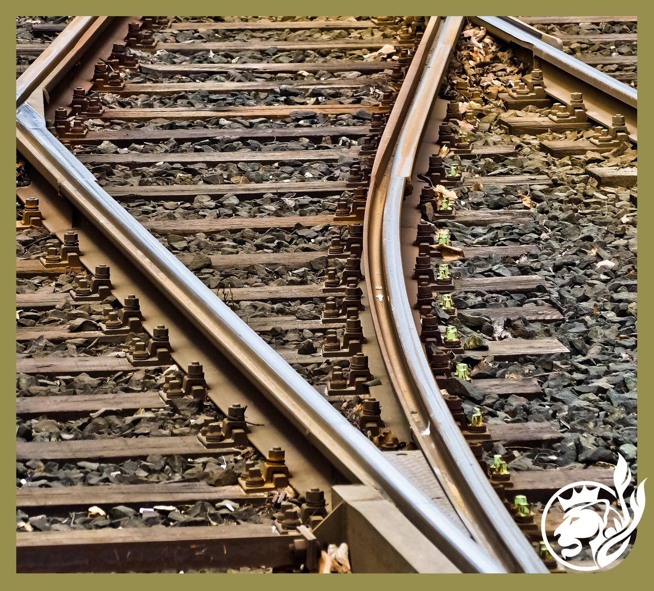 Nahaufnahme von Eisenbahnschienen mit Schotterunterlage und Stahlschienen, mit einem Konvergenzpunkt, verstreuten Steinen und grünen Pflanzen, ein goldenes Logo ist im unteren rechten Eck sichtbar.