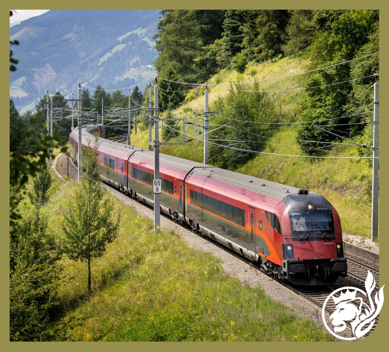 Ein roter Zug fährt auf einer Bahnstrecke durch eine hügelige Landschaft mit üppigem Grün und Bergen im Hintergrund.