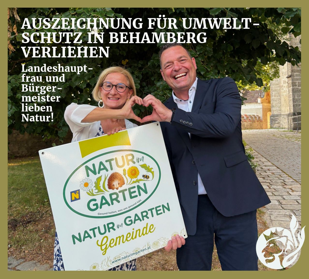 Ein Mann und eine Frau halten ein Schild für 'Natur im Garten' mit grünen und gelben Farben. Sie lächeln und bilden mit ihren Händen eine Herzform. Dahinter steht ein Baum mit grünen Blättern.