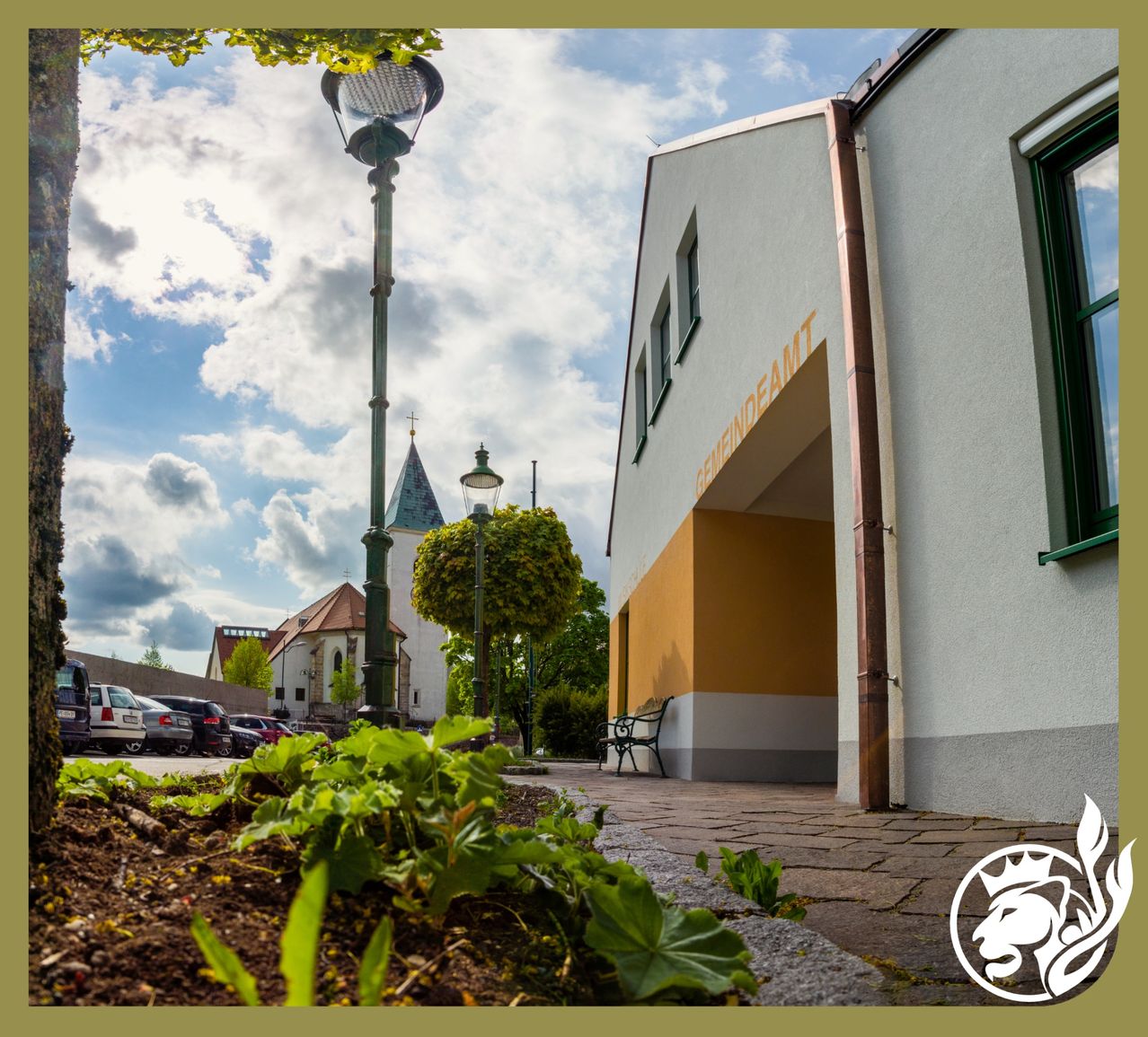 Ein Straßenblick auf ein Gebäude mit einem Namensschild, 'Reinendamt'. Eine Bank steht vor dem Eingang. Laternen stehen an den Seiten des Weges. Autos sind auf der linken Seite geparkt. Bäume stehen an den Straßenrändern.