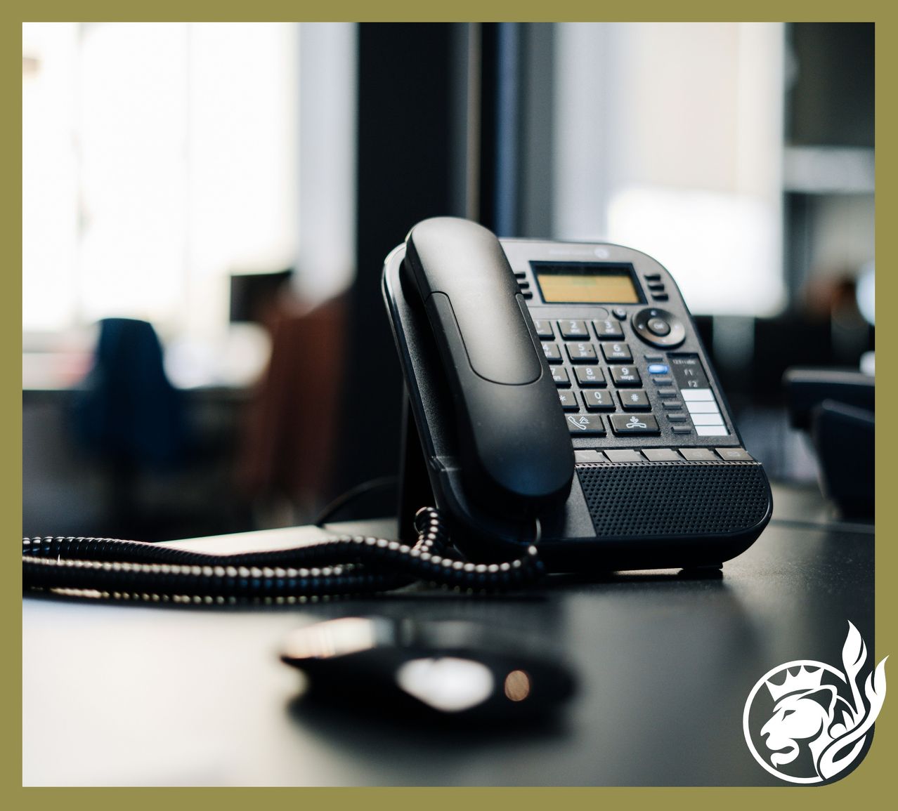 Ein schwarzes Telefon steht auf einem Schreibtisch neben einer Computermaus. Der Raum hat einen unscharfen Hintergrund mit Stühlen und einem Fenster.