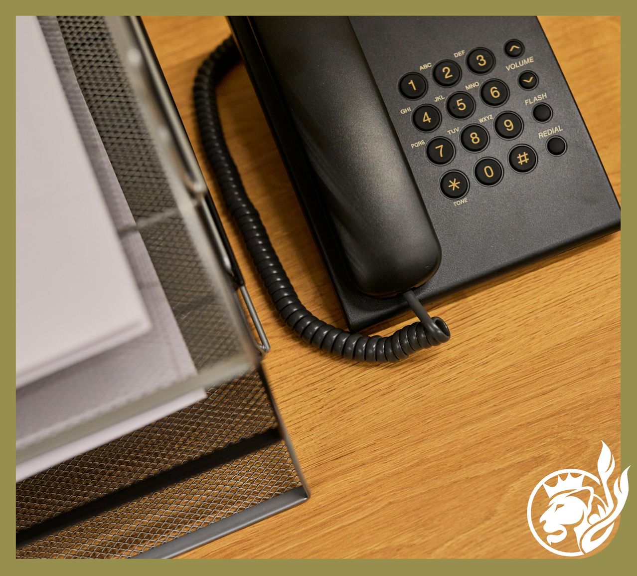 Ein schwarzer Telefonapparat mit goldenen Tasten steht auf einem hölzernen Schreibtisch neben einem klaren Glasorganisator.