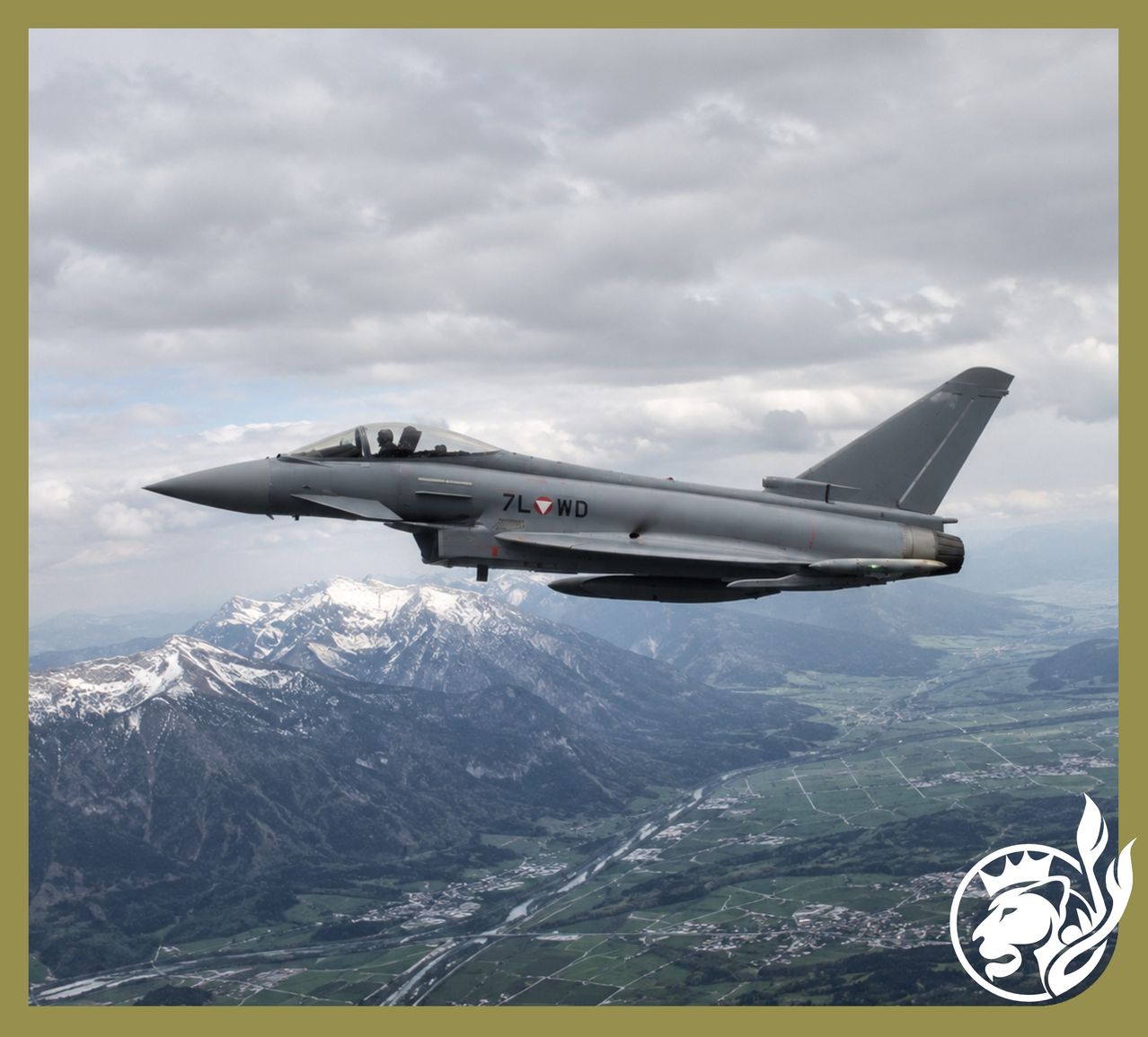 Ein Militärjet, gekennzeichnet mit 7L WD, fliegt über schneebedeckten Bergen unter einem bewölkten Himmel. Die Landschaft darunter zeigt Täler, einen Fluss und eine kleine Stadt.