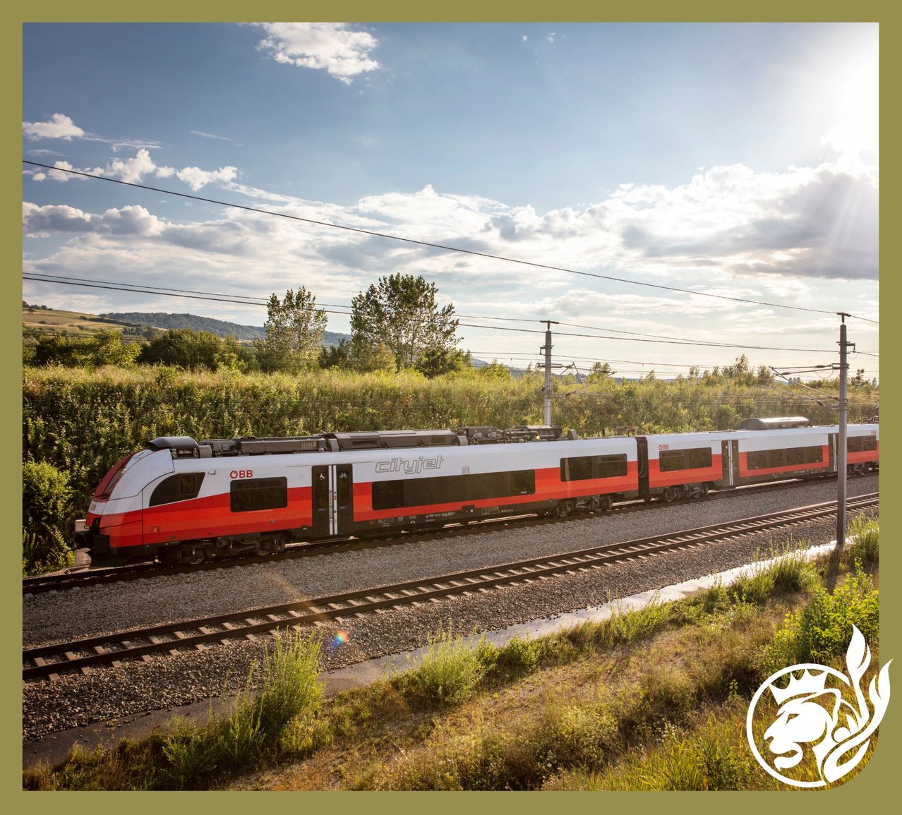Ein roter und weißer Zug mit CityJet Schriftzug bewegt sich auf der Bahnstrecke. Der Himmel ist klar mit einigen Wolken. Es gibt Bäume auf beiden Seiten der Strecke.