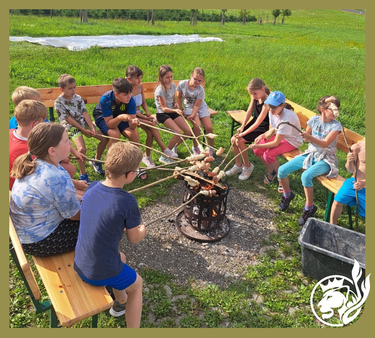 Eine Gruppe von Kindern sitzt um ein Feuer herum, jedes Kind hält einen Stock mit einem Marshmallow daran. Die Kinder lächeln und scheinen Spaß zu haben.