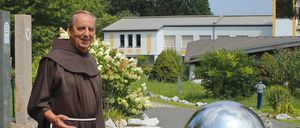 Ein Mönch in einer braunen Robe steht draußen vor einem Gebäude mit einem Kreuz auf der Spitze, umgeben von weißen Blumen und Bäumen.