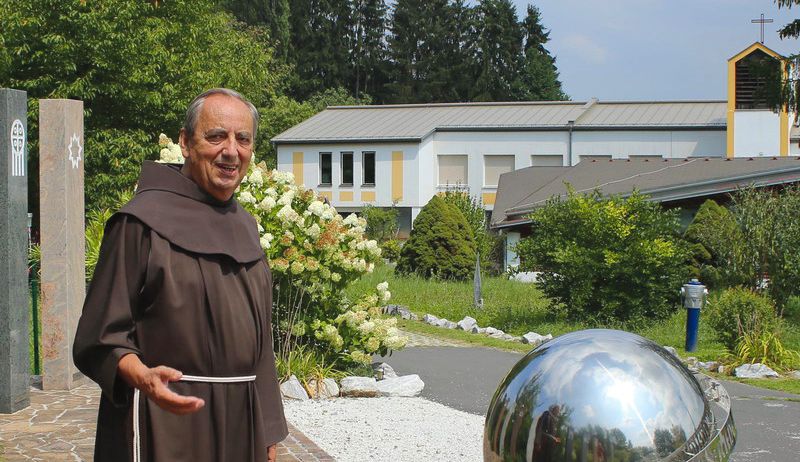 Ein Mönch in einer braunen Robe steht draußen vor einem Gebäude mit einem Kreuz auf der Spitze, umgeben von weißen Blumen und Bäumen.