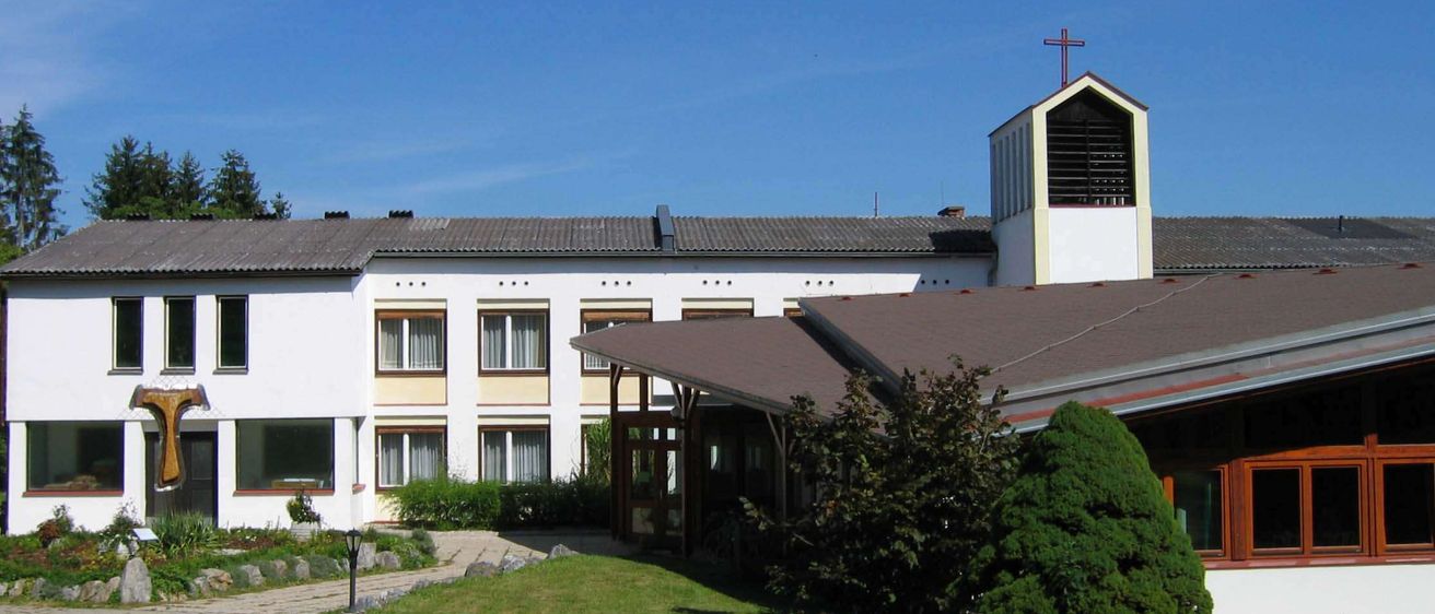 Ein weißes Gebäude mit einem braunen Dach und einem Kreuz darauf, umgeben von einem Rasen und Bäumen.