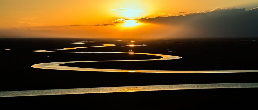 Sonnenuntergang über einem gewundenen Fluss, der Himmel in Orange- und Rosatönen gemalt, mit Wolken, die das Licht reflektieren.