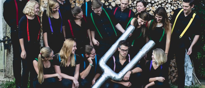 Ein Chor posiert für ein Foto vor einem Holzstapel, mit farbenfrohen Hosenträgern. Ein Mitglied hält ein großes silbernes Friedenszeichen.