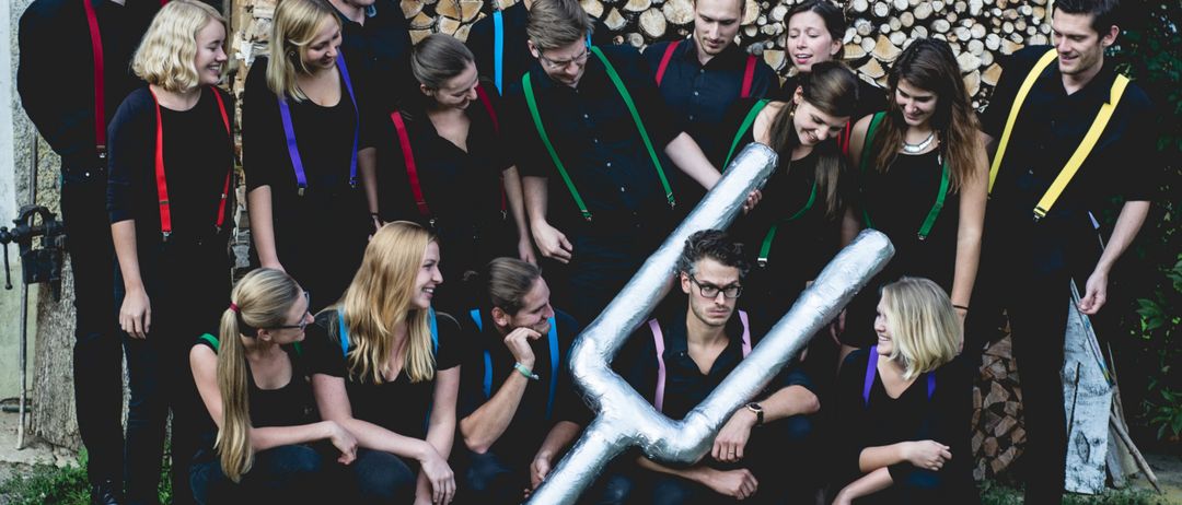 Ein Chor posiert für ein Foto vor einem Holzstapel, mit farbenfrohen Hosenträgern. Ein Mitglied hält ein großes silbernes Friedenszeichen.