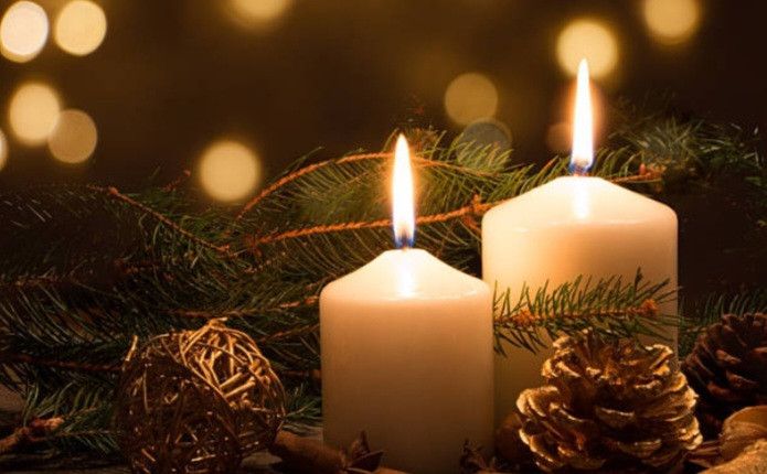 Zwei weiße brennende Kerzen vor einem Hintergrund aus immergrünen Pflanzen und Tannenzapfen.