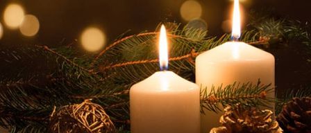 Zwei weiße brennende Kerzen vor einem Hintergrund aus immergrünen Pflanzen und Tannenzapfen.