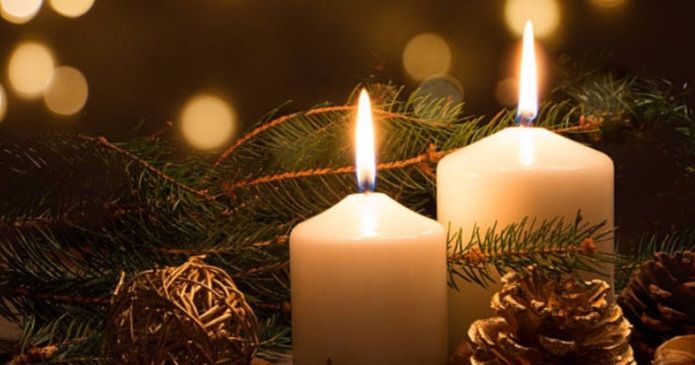 Zwei weiße brennende Kerzen vor einem Hintergrund aus immergrünen Pflanzen und Tannenzapfen.