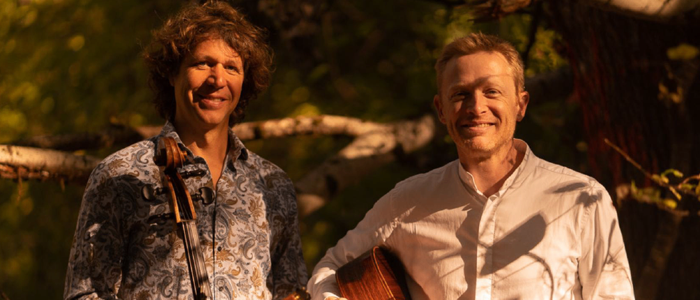 Zwei Männer lächeln und stehen nebeneinander, einer hält eine Geige und der andere eine Gitarre. Bäume und Äste im Hintergrund. Der Text lautet Egger, Gitarre Oskar Huetter, Violoncello.