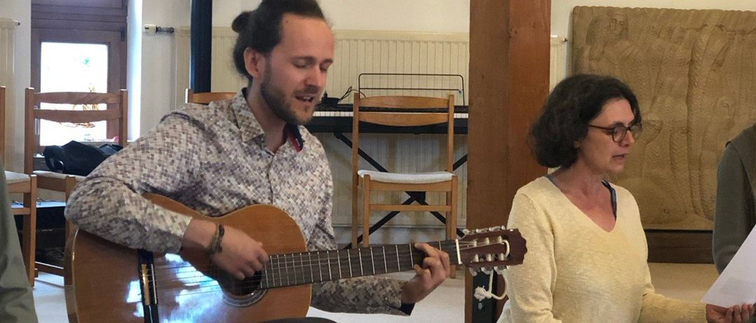 Ein Mann spielt Gitarre auf einer Bank, während eine Frau ein Papier liest, die auf dem Boden sitzt. Hinter ihnen stehen Stühle, ein Klavier und ein Kerzenhalter. Der Raum hat weiße Wände und Holzbalken.