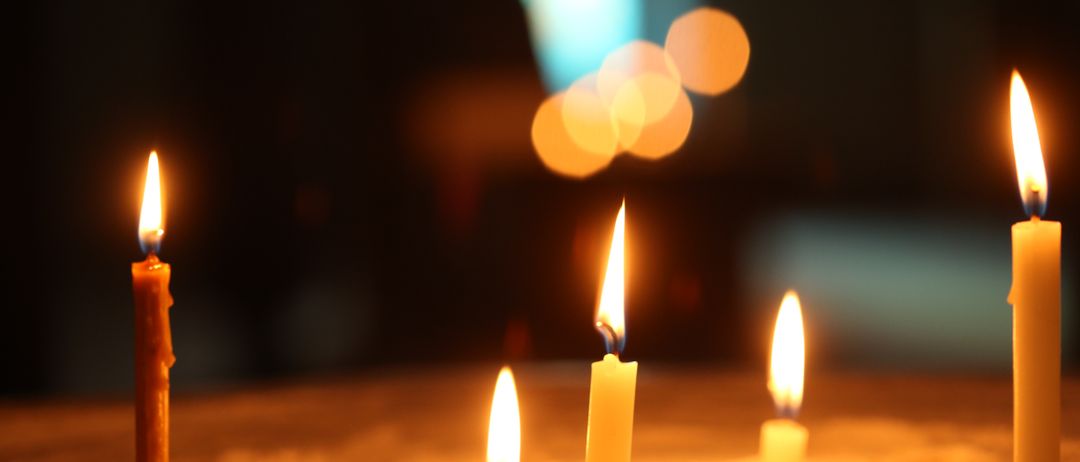 Drei Kerzen sind auf einer sandigen Oberfläche angezündet. Die mittlere Kerze ist vollständig beleuchtet, während die anderen teilweise beleuchtet sind. Der Hintergrund ist dunkel mit Bokeh-Lichtern.