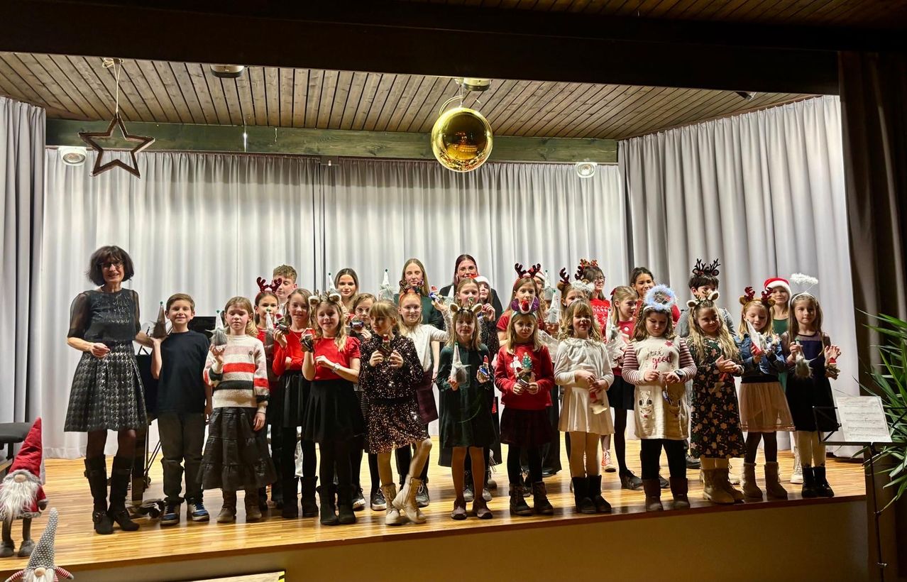 Eine Gruppe von Kindern und Erwachsenen in festlichen Kostümen steht auf der Bühne und hält verschiedene Gegenstände in den Händen. Sie führen wahrscheinlich ein Weihnachtsspiel auf.