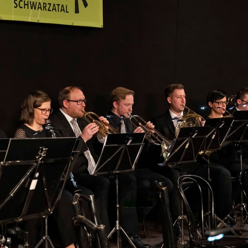 Eine Gruppe Musiker spielt Blechblasinstrumente auf der Bühne. Sie sind alle in formeller Kleidung gekleidet. Die Bühne hat einen schwarzen Hintergrund mit einem Banner, das 'Schwarztal' lautet.