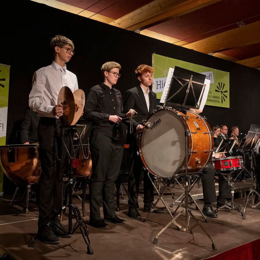 Eine Gruppe junger Musiker tritt auf der Bühne auf, wobei drei von ihnen Trommeln und Becken spielen, während andere verschiedene Instrumente im Hintergrund spielen.