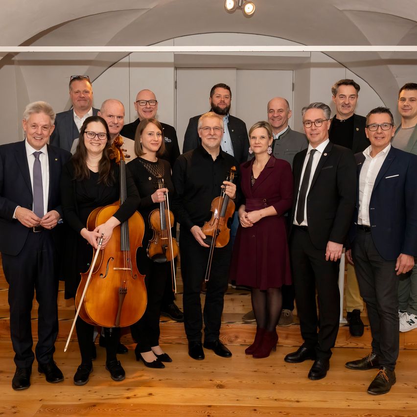 Eine Gruppe formell gekleideter Musiker steht im Kreis und hält verschiedene Streichinstrumente, wahrscheinlich in einem Studio oder Veranstaltungssaal.