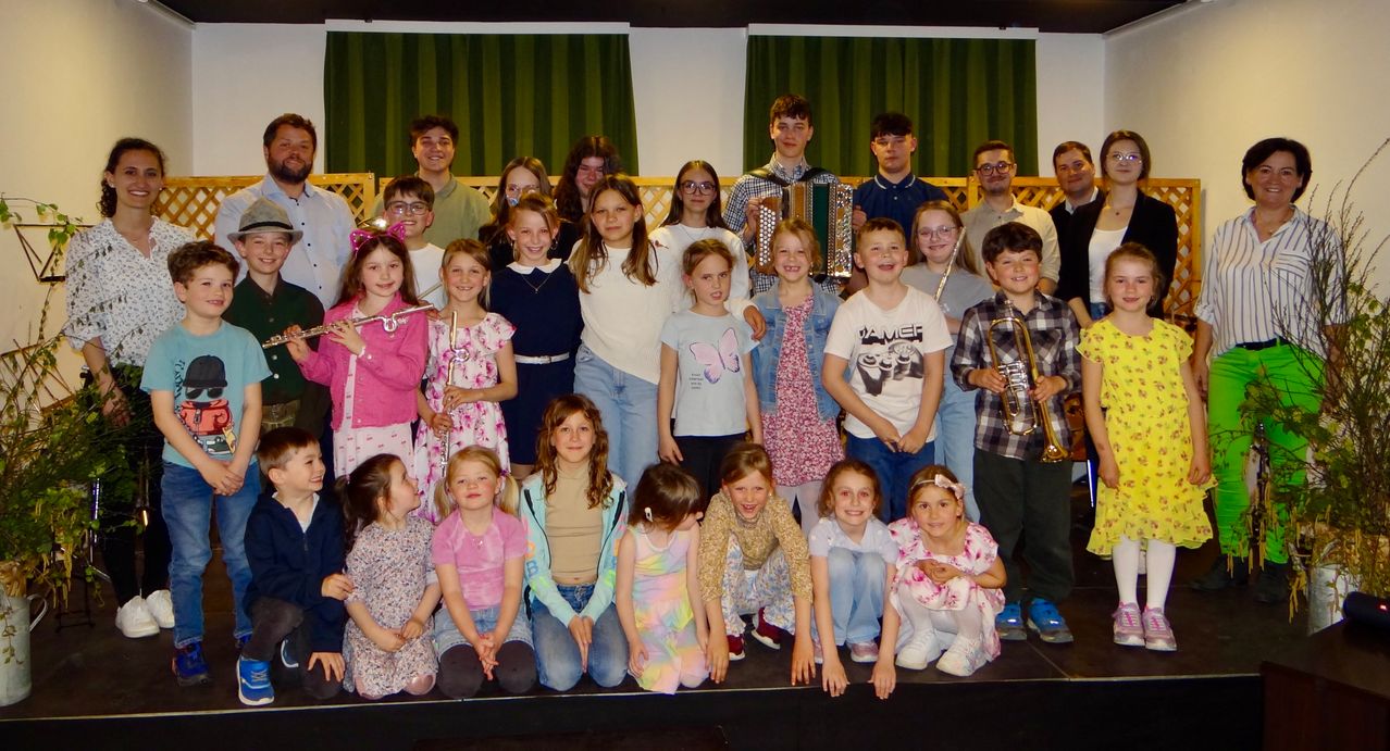 A group of children, both boys and girls, are standing on stage. Some are holding musical instruments like trumpets and flutes. They are smiling and posing for a group photo.