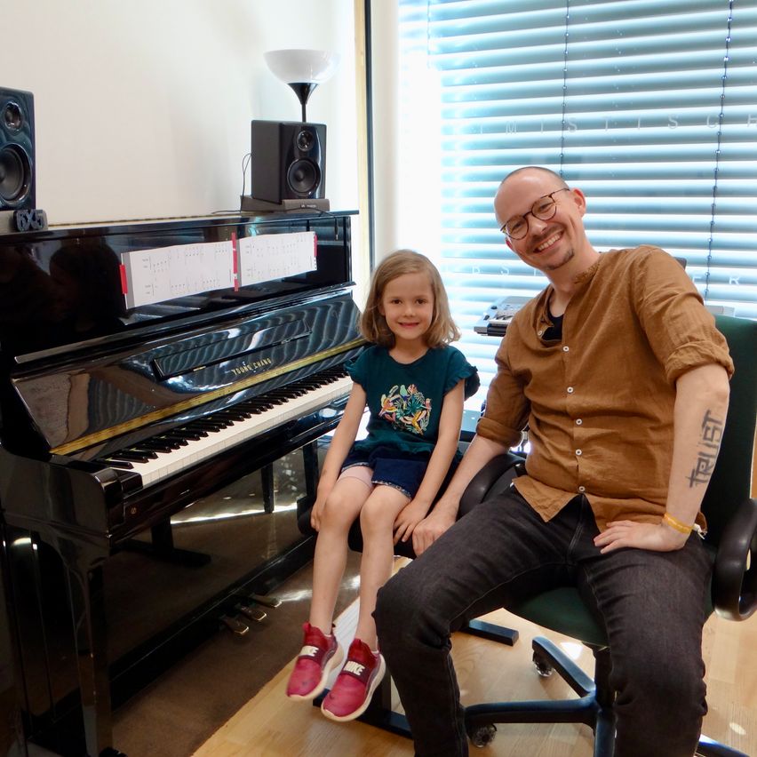 Ein Mann und ein kleines Mädchen sitzen zusammen am Klavier und lächeln für die Kamera. Auf dem Klavier liegen Musikblätter. Hinter ihnen stehen zwei Lautsprecher und eine Lampe.