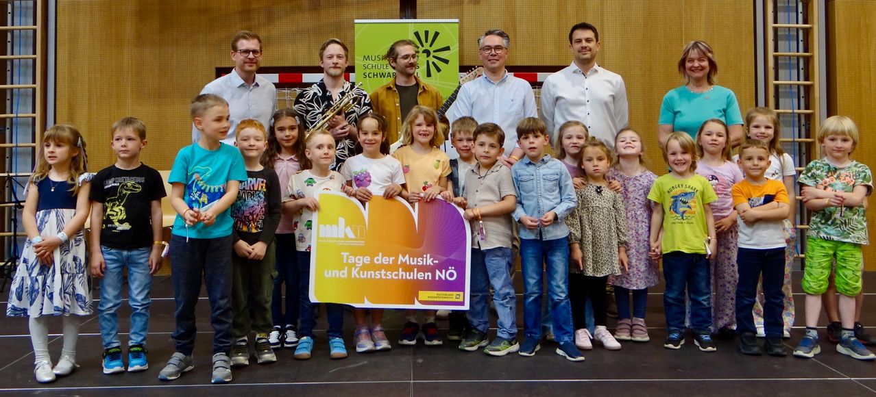 Eine Gruppe von Kindern und Erwachsenen posiert für ein Foto, einige halten Musikinstrumente, mit einem Banner für 'Tage der Musik- und Kunstschulen Nö'.