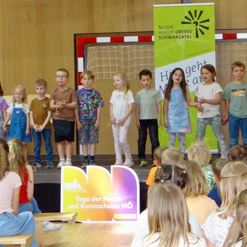 Kinder treten auf der Bühne auf, mit einem Banner im Hintergrund. Sie stehen in zwei Reihen. Die erste Reihe hält Händchen.