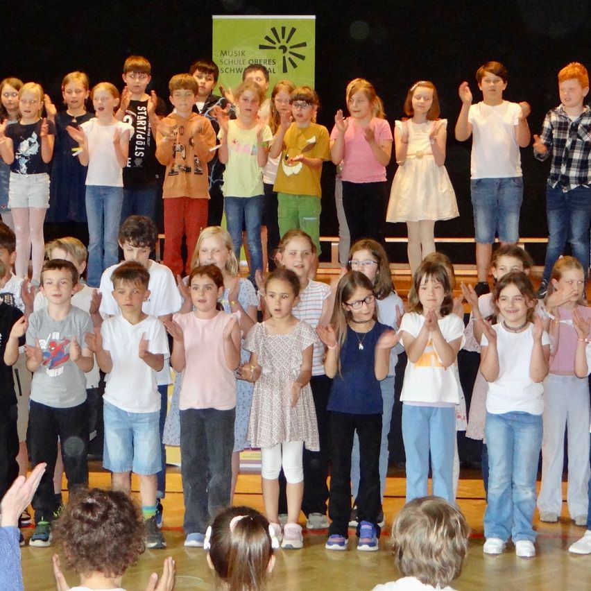 Eine Gruppe von Kindern klatscht auf der Bühne mit ihren Händen, trägt Freizeitkleidung. Einige tragen Brillen, Turnschuhe oder Armbänder. Ein grünes Banner ist über ihnen.