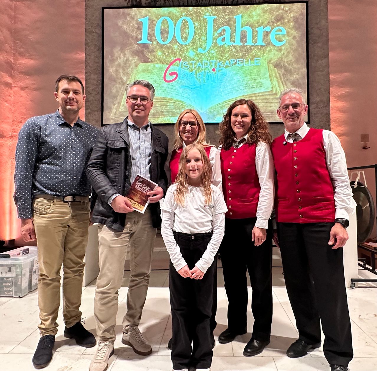 Fünf Erwachsene und ein junges Mädchen posieren vor einer Projektionsleinwand mit dem Text 100 Jahre Stadtkapelle. Das Mädchen hält ein Buch, und der Mann in der Mitte trägt eine Brille. Sie alle tragen formelle Kleidung, und eine große Glocke ist rechts sichtbar.