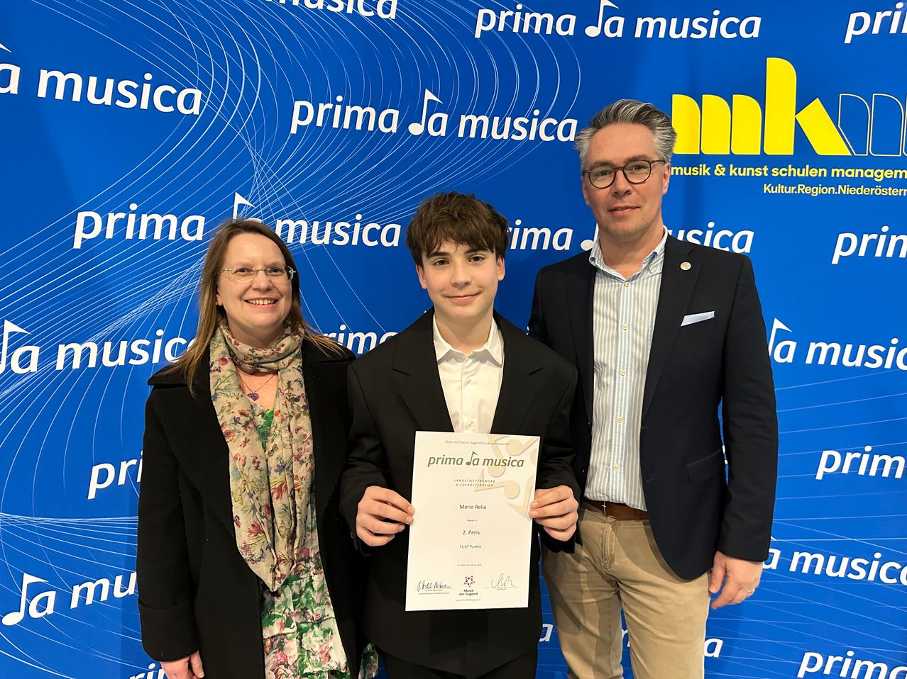 Drei Personen stehen vor einem blauen Hintergrund mit musikbezogenen Logos. Ein junger Junge hält ein Zertifikat, während eine Frau und ein Mann neben ihm stehen und lächeln.