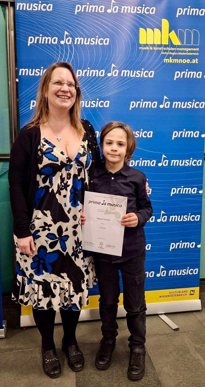 Eine Frau und ein junger Junge stehen lächelnd vor einem blauen Banner mit Noten und Text. Der Junge hält ein Zertifikat mit einer Note und Text, während die Frau ein Blumenkleid trägt.