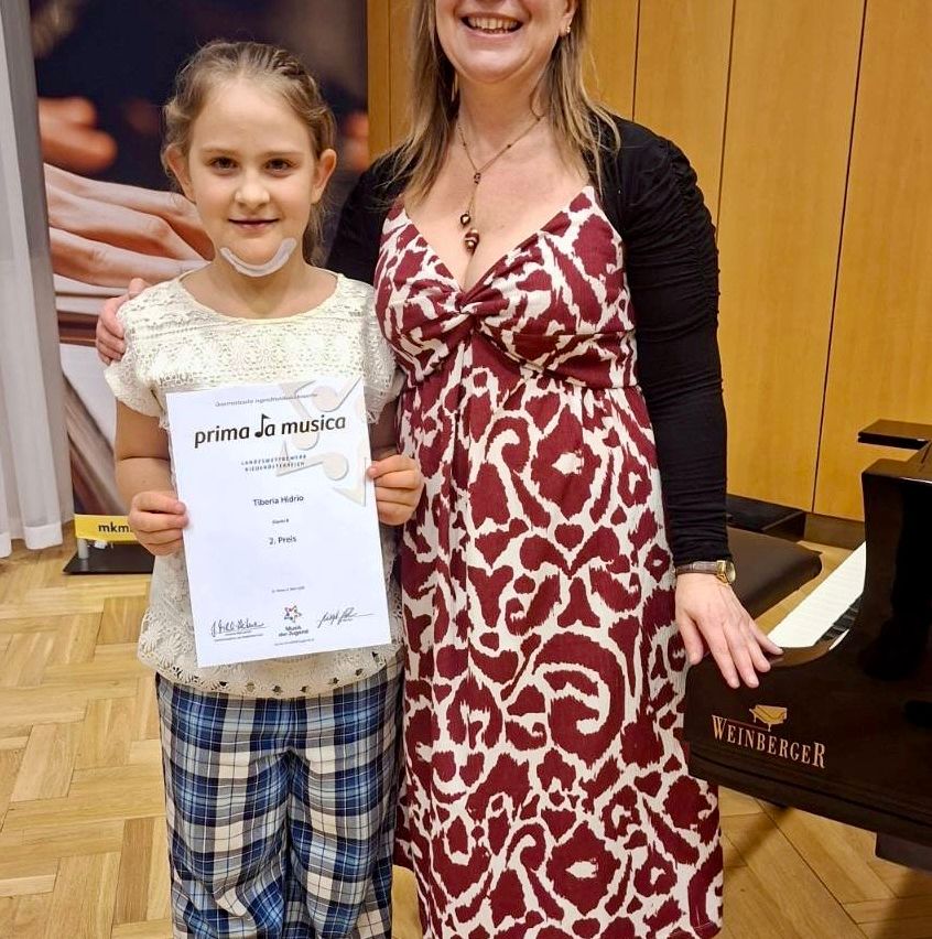 Ein junges Mädchen mit einem Zertifikat steht vor einem Klavier, mit einer lächelnden Frau hinter ihr. Sie befinden sich in einem Raum mit Holzboden und einer Holzwand.