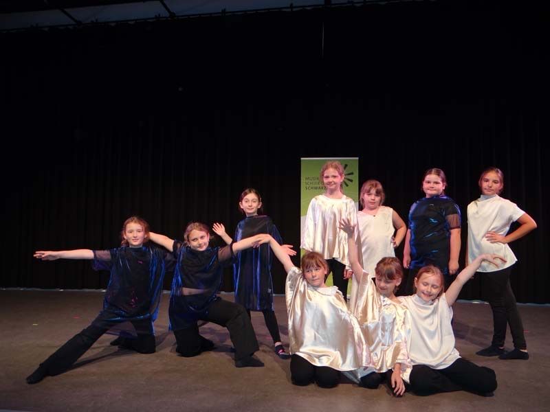 A group of young girls dressed in various colors are performing on a stage with a green backdrop. Some are kneeling while others stand and pose for a photograph.