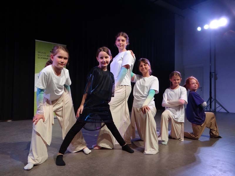 A group of young girls wearing white and black outfits are performing on stage, posing for a photo. Behind them is a green banner with text and a white door.