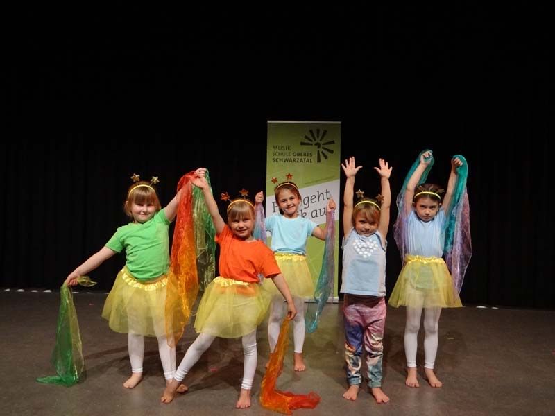 Five young girls in colorful costumes and headbands with stars are standing on a stage, possibly performing a dance. Behind them, a banner with German text is displayed.