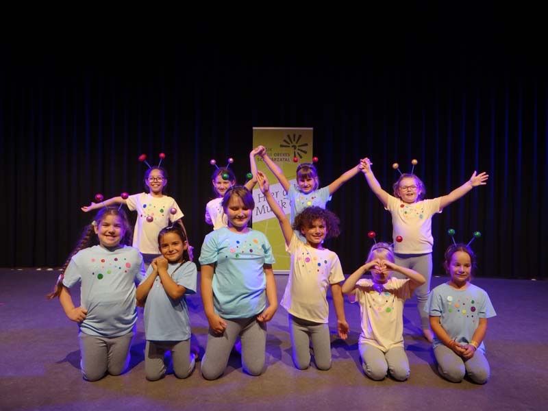 Group of young girls in matching outfits on a stage, some kneeling and some standing, all with headbands featuring antennae and colorful balls. Behind them is a banner with text.
