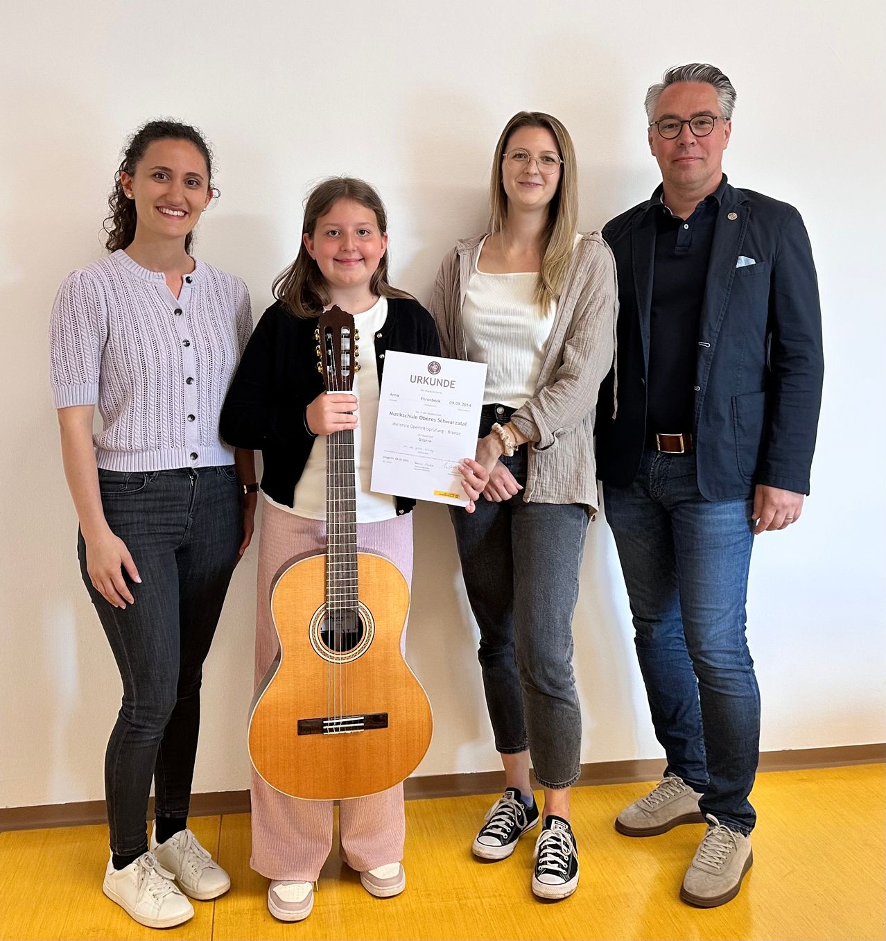 Vier Personen stehen lächelnd in einem Raum zusammen. Ein junges Mädchen hält ein Zertifikat und eine Gitarre. Eine Frau neben ihr hält das Zertifikat. Die anderen beiden sind Erwachsene. Sie stehen auf einem Holzboden.
