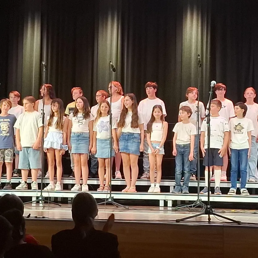 Ein Kinderchor in passenden weißen Shirts und Jeansshorts steht mit Mikrofonen auf der Bühne und tritt vor einem Publikum auf.