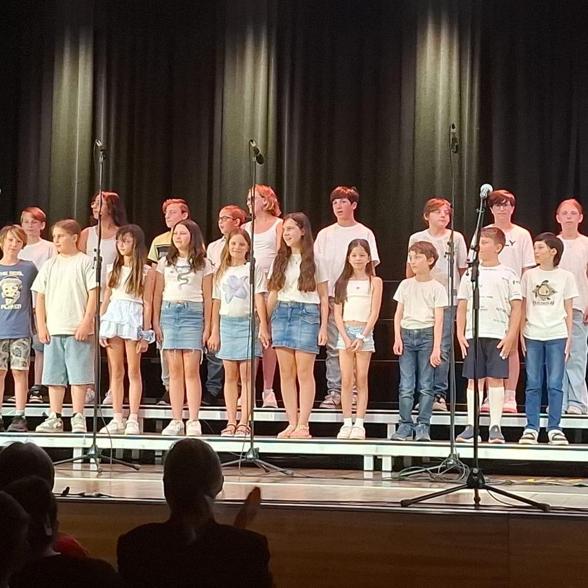 Ein Kinderchor in passenden weißen Shirts und Jeansshorts steht mit Mikrofonen auf der Bühne und tritt vor einem Publikum auf.