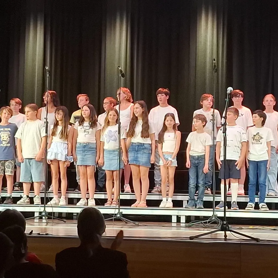 Ein Kinderchor in passenden weißen Shirts und Jeansshorts steht mit Mikrofonen auf der Bühne und tritt vor einem Publikum auf.
